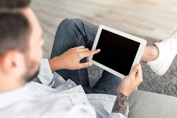 Young man using digital tablet with blank black screen, browsing website or showing new app, sitting on sofa at home. Advertisement concept. Device with empty screen, mockup for your design