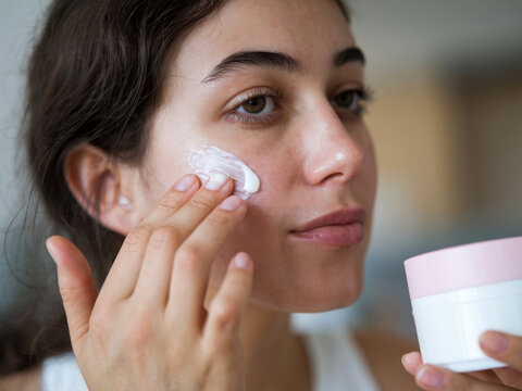 Un primer plano de una mujer joven con cabello oscuro aplic&aacute;ndose crema hidratante en la cara. Ella est&aacute; usando sus dedos para esparcir la crema sobre su piel
