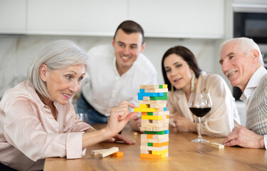 Joyful senior parents and young couple sitting around table playing brick game and drinking wine in goblets at home