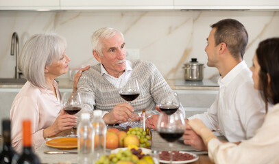 Happy senior man celebrating birthday with wife and adult children over cozy family dinner table served with wine and light snacks..