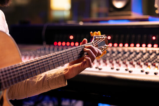Skilled singer performing on his electro acoustic guitar in control room, recording new sounds and mixing with audio console. Musician and technician composing a hit song for an album. Close up.