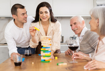 Group of mature spouses and middle-aged male visitor with girlfriend is playing board game. Senior married couple build tower with wooden blocks bricks with younger spouses