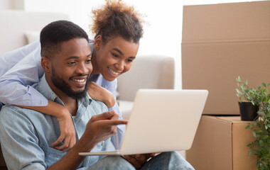 Fototapeta premium Smiling Black Spouses Using Laptop Sitting On Sofa Among Packed Boxes In New Home. Moving House Concept. Selective Focus