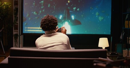 African american gamer playing classic arcade space shooter videogame on big smart TV, shooting asteroids. Zoom in shot on man using gaming console to solve missions in singleplayer game © DC Studio
