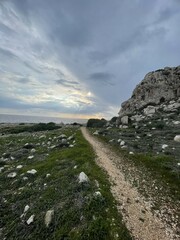 Serene Coastal Pathway with Dramatic Rocky Cliff and Moody Overcast Sky Leading to a Sunset Horizon Over the Sea