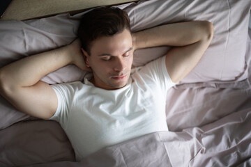 Man resting peacefully in bed with hands behind his head, eyes closed, wearing a white t-shirt, surrounded by pastel bedding, creating a serene and cozy atmosphere in soft natural light.