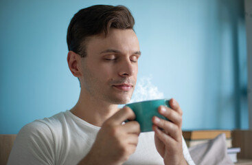 Close-up of a relaxed man in a white t-shirt holding a steaming turquoise cup, eyes closed,...