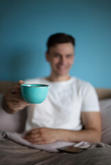 Blurred smiling man in a white t-shirt sitting on a bed, offering a turquoise cup towards the camera, with pastel bedding and a light blue background, creating a warm and inviting atmosphere.