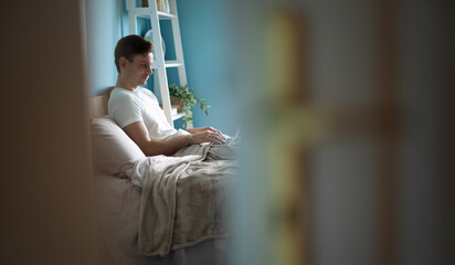 A man in a white t-shirt sits on a bed, working on a laptop in a cozy bedroom with blue walls, plants, and soft natural light, captured from a unique doorway perspective.