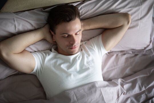 Man resting peacefully in bed with hands behind his head, eyes closed, wearing a white t-shirt, surrounded by pastel bedding, creating a serene and cozy atmosphere in soft natural light.