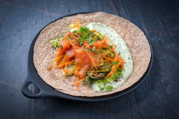Italian style wrap piada tortilla with cured graved salmon, salad and zucchini served as close-up with avocado sour cream dressing dip on a Nordic Design plate