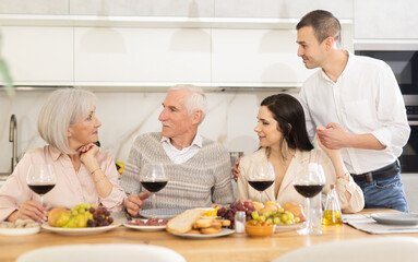 Merry old family members having lunch with their son and his girlfriend while drinking wine and talking warmly in their house