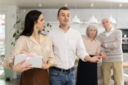 Positive interested young adult man gesturing welcomingly towards female guest holding gift box, against backdrop of disapproving elderly parents in domestic setting.... - Powered by Adobe