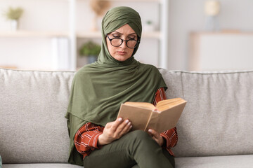 Mature Muslim Female Reading Book Wearing Eyeglasses Sitting On Sofa At Home Relaxing On Weekend....
