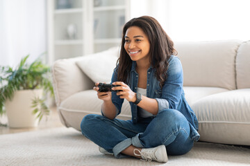 A young woman sits comfortably on the floor, playing video games with a joyful expression. The cozy living room features a modern sofa and indoor plants, creating a relaxing atmosphere.