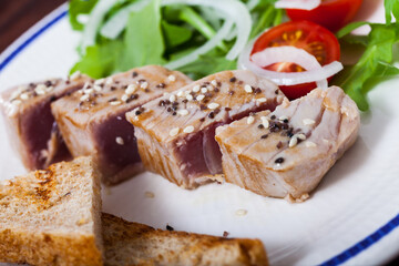 Delicious slightly fried tuna sprinkled with sesame served on plate with toasts, fresh tomatoes and greens