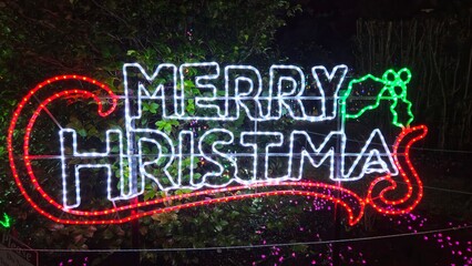 A bright "Merry Christmas" sign made of white, red, and green LED lights glows against a backdrop of bushes at night, with holly leaves and candy cane shapes enhancing the festive display.