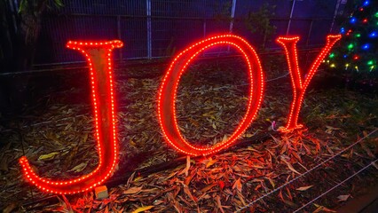 A large "JOY" sign outlined with bright red LED lights at night, set against a background of fallen leaves and a faintly visible Christmas tree with multicolored lights.