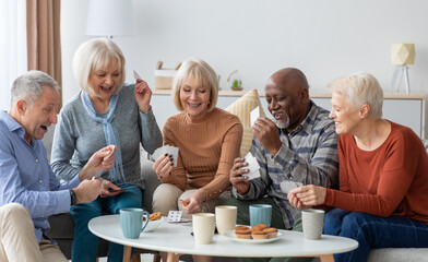 Multiethnic group of happy senior friends men and women in casual sitting on couch, having conversation and playing cards while drinking tea with cookies, chilling together at home