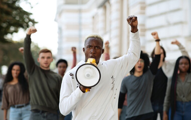 African american man with megaphone leading international group of protestors on the street, raising fist up, chanting slogans. Leader of strikers with loudspeaker yelling demands to government