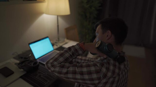 Cervical neck ache, nerve arthritis. Man studying late through night at laptop computer with cervical pain. Exhausted guy relaxing his neck using gun massager while overworking at notebook computer. 