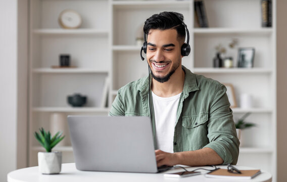 Happy handsome young islamic man with beard in headphones looks at laptop in living room interior. Consultation remotely, social distance, new normal and customer support during covid-19 quarantine