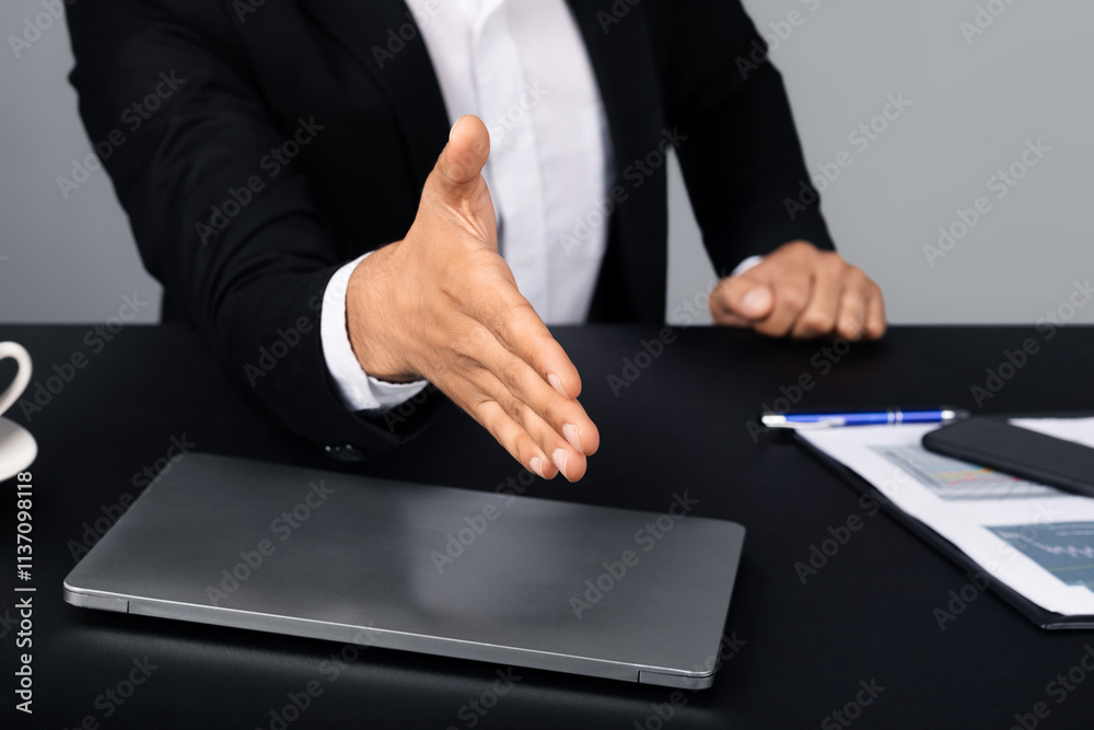 Poster Cropped of entrepreneur in a suit offers a handshake across a desk with a laptop and documents. The setting reflects a modern corporate environment, emphasizing professional interaction.