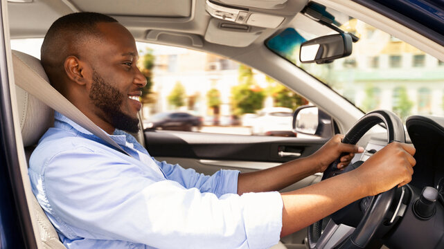 Side view profile portrait of joyful African American man driving car, going to work or on trip during summer vacation. Happy guy riding his brand new nice car, doing test drive in dealership showroom