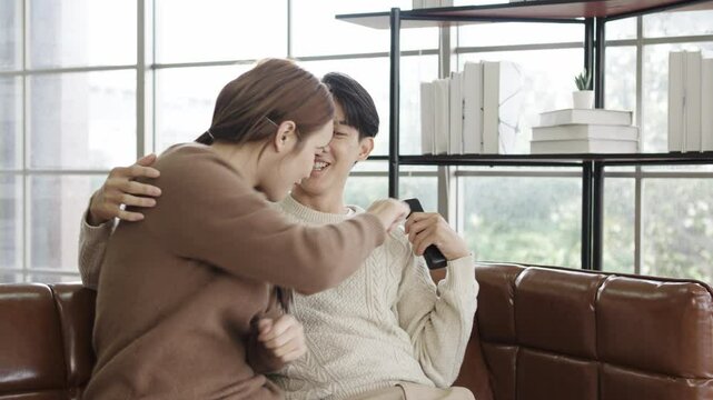 Happy young asian couple with a remote control for choice and relax. 
playing and trying to change the channel, playful and teasing while watching television in the living room at home.