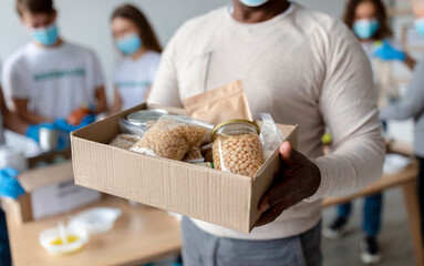 Senior black man holding box with donations food, cans and packages with grains and pasta, cropped. Group of millennial volunteers packing boxes with products for poor and homeless people