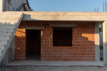 Unfinished residential building with red brick walls and concrete staircase under construction in sunlight. Concept of architecture, construction process, and structural development in urban areas. 