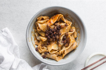 Overhead view of spicy wide noodles in a grey bowl, top view of biangbiang mian in a bowl, spicy belt noodles with green onions and diced pork in a bowl