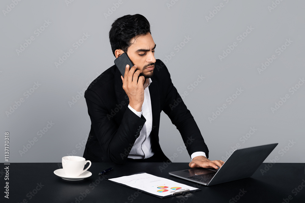 Poster A Middle Eastern businessman is focused on a phone call while typing on his laptop in a modern studio.