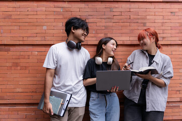 Three young students collaborating on a project, using laptop and notebook, exchanging ideas and knowledge outdoors against a brick wall