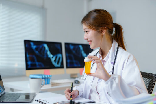 Asian female doctor holding medicine bottle and taking notes while having online consultation and working with dna double helix structure on computer screen