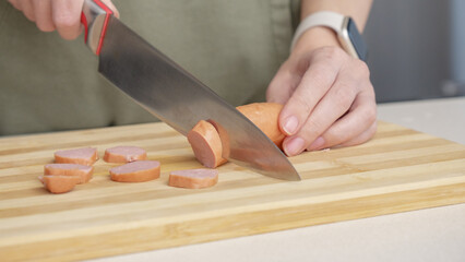 Close-up of a woman cutting sausage on a wooden cutting board in a kitchen setting with different ingredients.