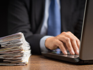 Close-up of a businessman working on a laptop with a tall stack of paperwork on the desk.