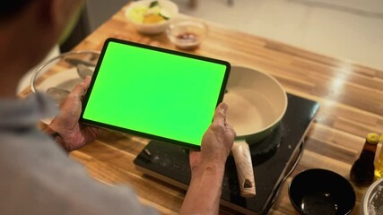 Senior man using digital tablet with a blank green screen while cooking in his home kitchen