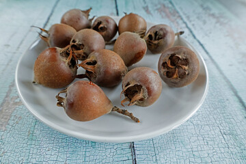 A plate of ripe medlar fruits arranged on a white dish, set against a rustic wooden table. The fruits have a brownish hue and a slightly wrinkled texture, typical of their ripened state.