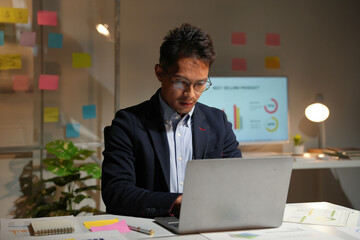 Asian businessman concentrating on his work on a laptop in a modern office late at night, surrounded by sticky notes and charts