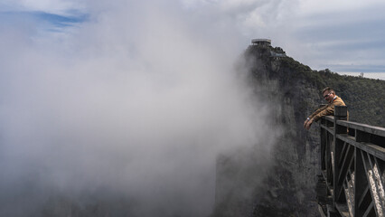 The man is standing on the observation deck, leaning on the lattice railing, smiling. In the distance is a mountain with a survey platform, shrouded in clouds. China. Tianmen Mountain. Zhangjiajie.