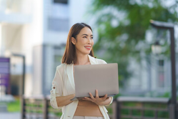 Asian businesswoman smiling and holding laptop while standing in a green urban business district