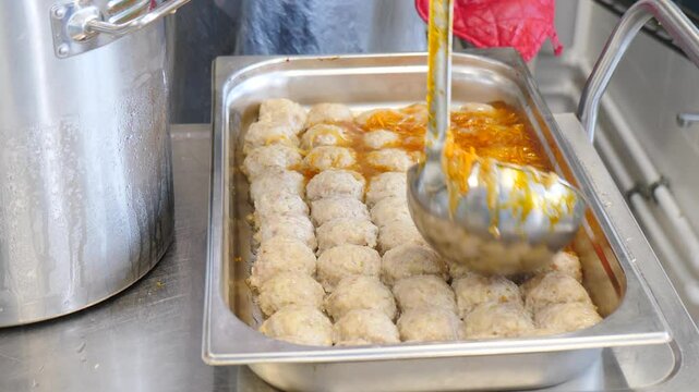 Chef is pouring tomato sauce on a tray of meatballs in a professional kitchen. The sauce is bright red and the meatballs are arranged in neat rows. 4 k video