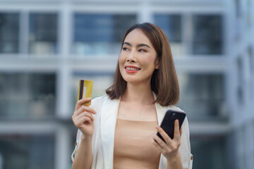 Businesswoman holding a credit card and using a smartphone, enjoying shopping online in front of a modern building