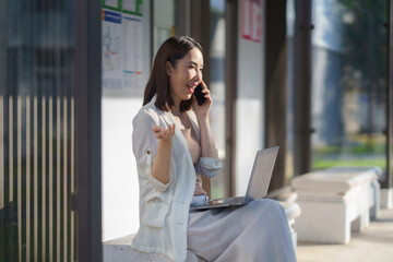 Fototapeta premium Asian businesswoman using laptop and talking on mobile phone while sitting on bench outdoors