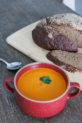 Freshly made tomato soup in a red ceramic bowl on a wooden dining table with a fresh loaf of rye bread on a wooden chopping board