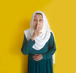 Keep Silence. Elderly Asian Muslim Woman Putting Finger To Her Mouth And Standing Against Yellow Background