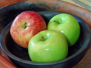 Two green apples and one red apple resting in black clay bowl