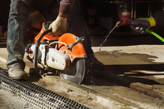 Worker operates a cutting machine on wet concrete while another sprays water to control dust at a construction site.
