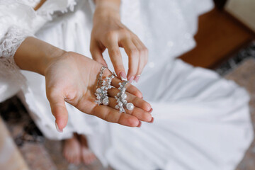 A woman is holding a pair of earrings in her hand. The earrings are made of pearls and are very shiny. The woman is wearing a white dress and is sitting on a couch. The scene is very elegant